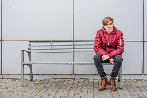 Teenager boy sitting on metal bench outdoor in city over urban gray background waiting