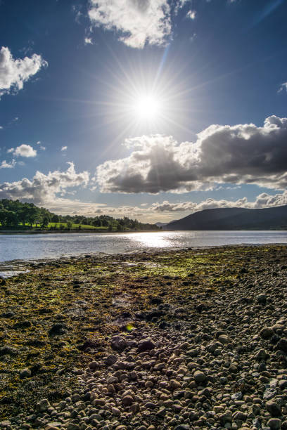The sun in a blue sky over Loch Linnhe with a pebble beach