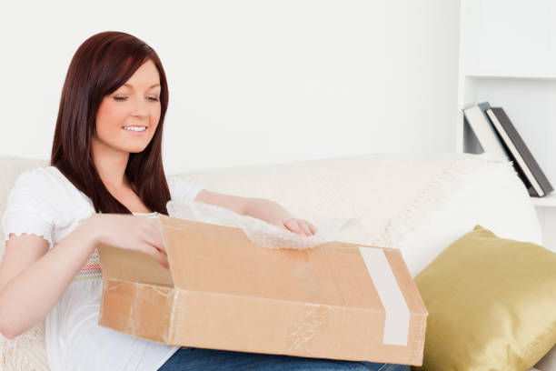 istock Attractive redhaired woman opening a carboard box while sitting on a sofa 827197868