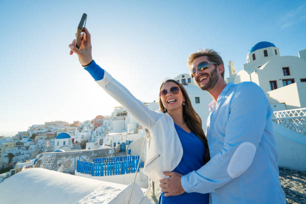 City Breaks, smiling couple taking selfie in santorini couple on their honeymoon greek island santorini taking a selfie with their smartphone in front of the typical architectre in greece with chapels and white houses background happy couple on vacation in santorini greece stock pictures, royalty-free photos & images