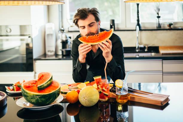 красивый мужчина делает фруктовый салат и ест арбуз на кухне - fruit salad fruit healthy eating making стоковые фото и изображения
