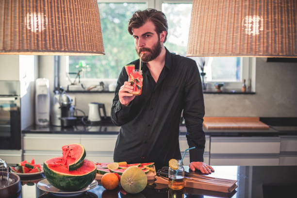 красивый бородатый мужчина пьет арбузный сок и делает фруктовый салат на кухне - fruit salad fruit healthy eating making стоковые фото и изображения