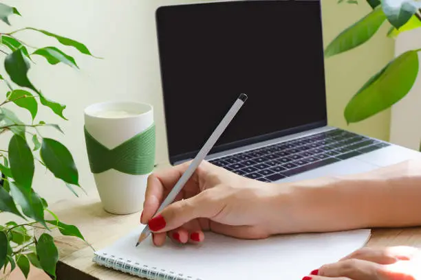A woman with bright manicure writes in pencil in the diary. Beside to the table is a laptop, a cup of water with lemon and green plants. The concept of a greened workplace or office.