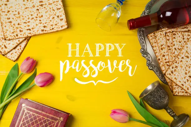Jewish holiday Passover Pesah celebration with matzoh, tulip flowers and wine bottle on yellow wooden background. View from above
