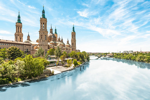 Scenery landscape with Basilica of Our Lady of Pillar. Blue sky reflects in clear water. Green trees along river. Famous church with beautiful architecture. Famous tourist place in Spain, Europe.
