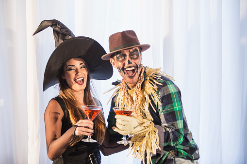 A couple in their 30s having fun at an adult halloween party, drinking cocktails, laughing at the camera. The woman is wearing a witch costume and the man is a scarecrow.