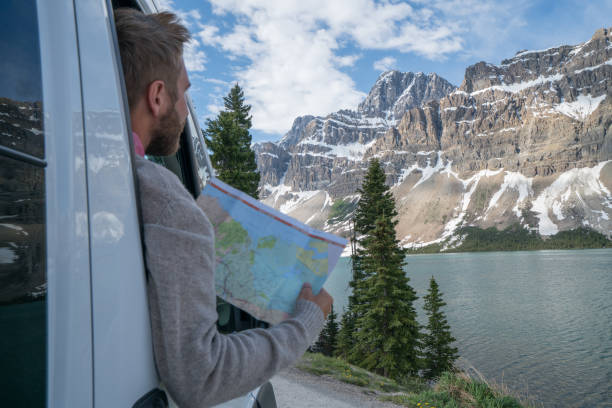 Young man looks at road map near mountain lake Young man in car on mountain road looks at map for directions. Mountain lake landscape in Springtime with snow melting. road map of canada stock pictures, royalty-free photos & images