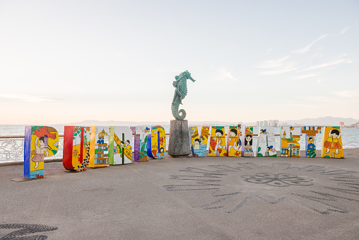 This is a horizontal, color photograph shot in travel destination, Puerto Vallarta, Mexico. Along the malecon are very large 3D letters that spell out Puerto Vallarta. A sea horse sculpture is placed in the middle. The sunset backlights the scene.