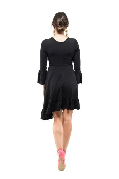 Back view of young woman with bun hairstyle and black flounced dress walking away. Full body length portrait isolated on white studio background.