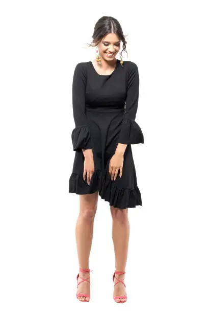 Joyful laughing young woman in black dress looking down with windy flying clothes. Full body length portrait isolated on white studio background.