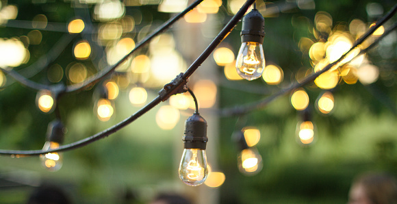 Strings of lights are set above a summer picnic gathering. The lights hang from wires to decorate and illuminate the space where the guests below are dining.