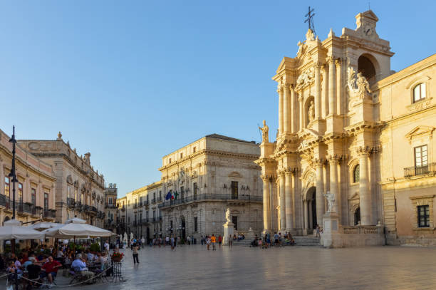 The Piazza Duomo and cathedral on the island of Ortiga in Syracuse, Sicily, Italy. Photo taken during sunset, and shows the main square with some visitors seeking dinner at one of nearby restaurants.