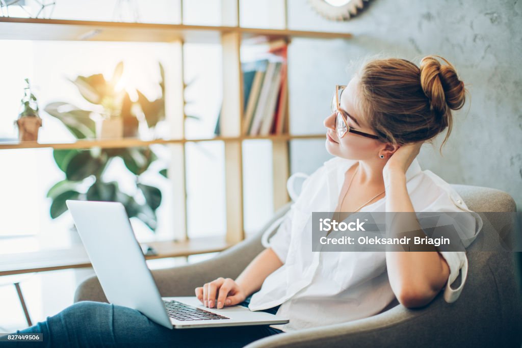Young beautiful woman working in an alternative office Young beautiful woman in her 20s wearing glasses working with a laptop while sitting in a chair in an alternative office. Change Stock Photo