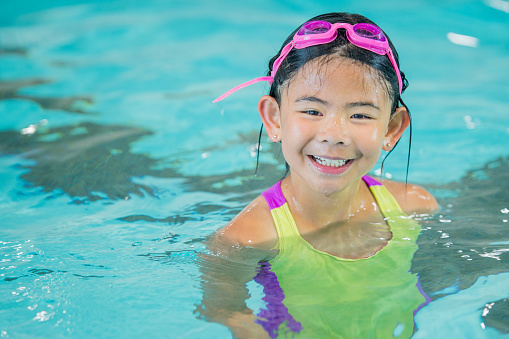 An Asian elementary school girl is indoors in a swimming pool. She is wearing a swimsuit and goggles. She is smiling at the camera while swimming.