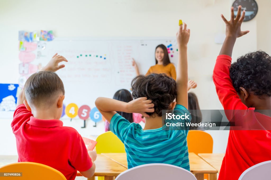 Bambino in età prescolare alzare il braccio per rispondere alla domanda degli insegnanti sulla lavagna in classe, concetto di educazione all'asilo - Foto stock royalty-free di Aula