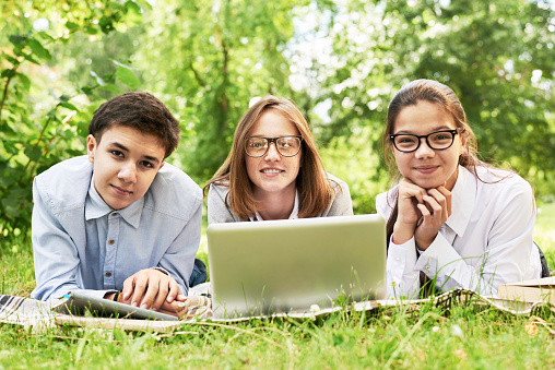 Three smiling school children enjoying picnic on green lawn using laptop and looking at camera