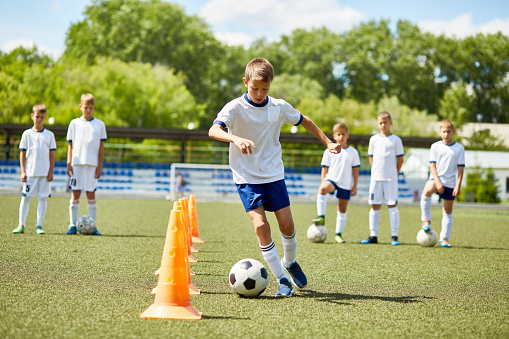 Portrait of boy in junior football team  leading ball between cones during  practice in field on sunny day