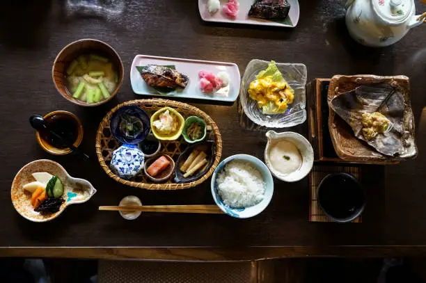 Japanese ryokan breakfast dishes including cooked white rice, grilled fish, fried egg, soup, mentaiko, pickle, seaweed, hot plate, other side dishes and green tea on wooden table, Japan