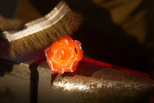 Blacksmith is descaling the red-hot metal flower with the strip brush for cleaning metal. Shooting at dark blacksmith workshop