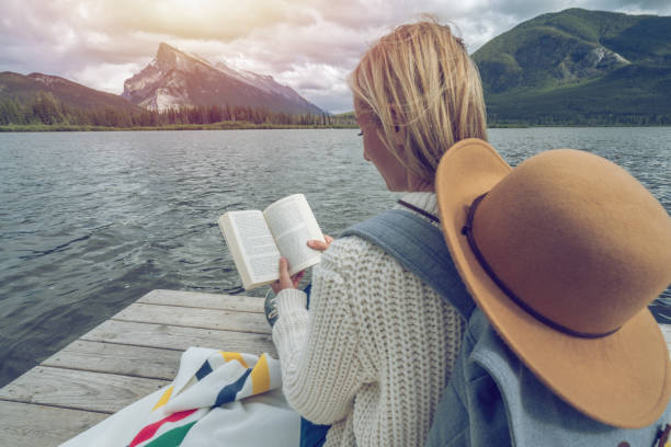 istock Young woman reading book on lake pier 827535910
