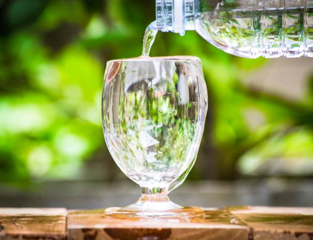 istock Glass water on wood board with tree blurry bokeh background. using wallpaper or background for drink photo. Take of the product. 827540520