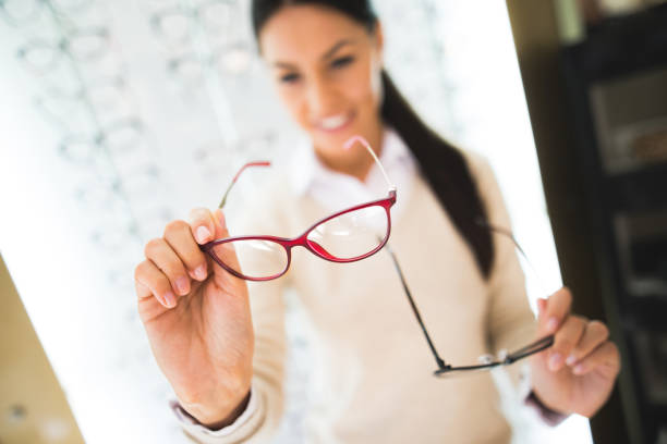 Blurred image of a young smiling woman selecting eyeglasses in optical shop. Focus on eyewear in the foreground.
