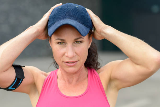 Cute attractive young woman in a baseball cap wearing a strap on her arm for her mobile phone looking intently at the camera with a quiet smile