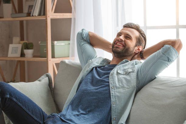 jeune homme assis sur un entraîneur reste à la maison - bachelor home photos et images de collection