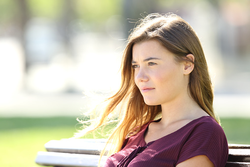 Portrait of a serious girl sitting on a bench thinking and looking away outdoors