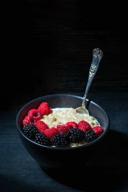 High resolution digital caputure of a bowl of overnight oats with fresh raspberries and blackberries and a splash of cream. Oats are in a black bowl, sitting on a black weathered surface, and the lighting is subdued and omnidirectional. There is an ornate silver spoon plunged partially into the oats. Some steam is visible, rising from the surface of the oats. There is ample text space in the upper half of the frame.