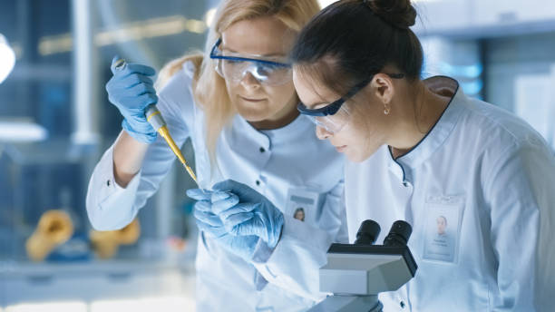 medical research scientist gouttes échantillon sur la lame et sa collègue examine il sous microscope. ils travaillent dans un laboratoire moderne. - drug discovery photos et images de collection