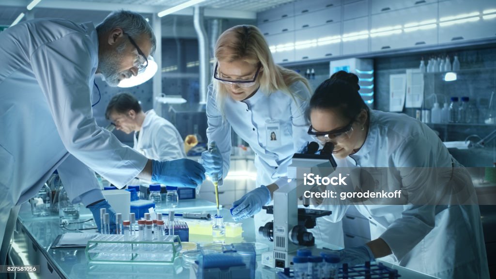 Team of Medical Research Scientists Work on a New Generation Disease Cure. They use Microscope, Test Tubes, Micropipette and Writing Down Analysis Results. Laboratory Looks Busy, Bright and Modern. Scientist Stock Photo