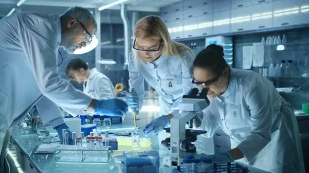 Photo of Team of Medical Research Scientists Work on a New Generation Disease Cure. They use Microscope, Test Tubes, Micropipette and Writing Down Analysis Results. Laboratory Looks Busy, Bright and Modern.