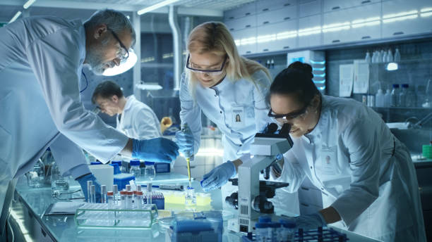 team of medical research scientists work on a new generation disease cure. they use microscope, test tubes, micropipette and writing down analysis results. laboratory looks busy, bright and modern. - lab imagens e fotografias de stock