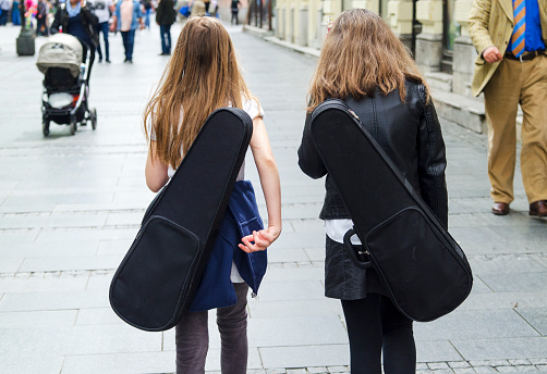 The back of two little girl friend walk together on the street with violin in box.