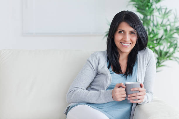 istock Smiling woman holding a mug with two hands while resting on a sofa 827646256