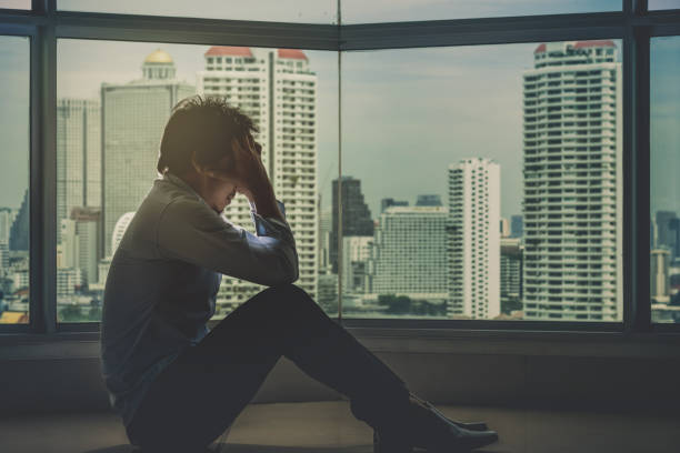 istock depressed man sitting head in hands on the interior Skyscraper with low light environment beside the windows over the cityscape background, dramatic concept 827647542
