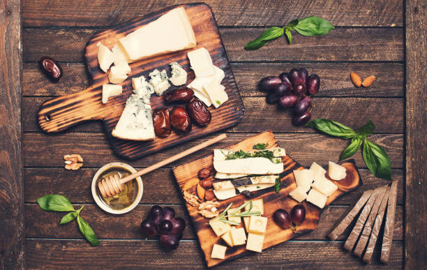 istock Assorted cheeses on wooden board plates served with nuts, grapes, bread, honey, and dates on dark wood background, top view. 827650824