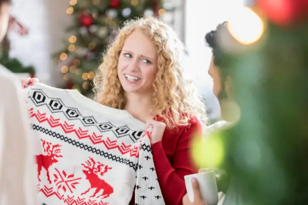 Young Caucasian woman makes a face while holding an ugly Christmas sweater. She and her friends are participating in an ugly Christmas sweater gift exchange.