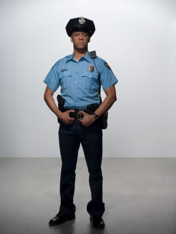 Confident police officer in black uniform posing with weapon indoors. Front view of bearded policeman with hand on belt, holding police handgun, isolated on white. Concept of work, profession, weapon.