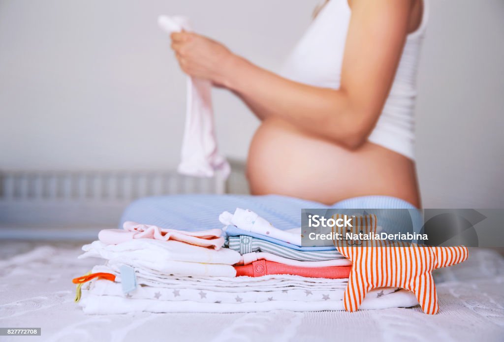Pile of baby clothes, necessities and pregnant woman on bed in home interior of bedroom. Pile of baby clothes, necessities and pregnant woman on bed in home interior of bedroom. Pregnant woman is getting ready for the maternity hospital, packing baby stuff. Pregnancy, birth concept. Pregnant Stock Photo