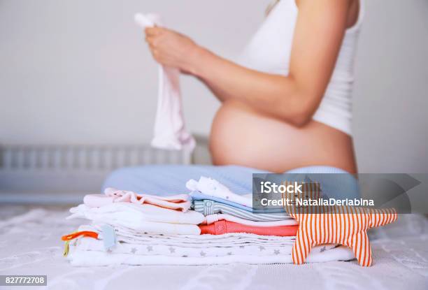 Pile Of Baby Clothes Necessities And Pregnant Woman On Bed In Home Interior Of Bedroom Stock Photo - Download Image Now