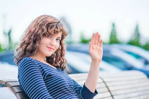 Portrait of young brunette woman wave gesturing while sitting on bench in outdoor park