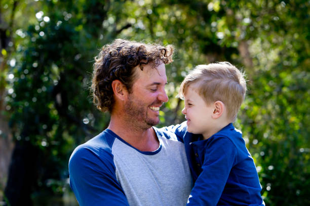 Father holding young son outdoors in sunlight in park - looking away from camera