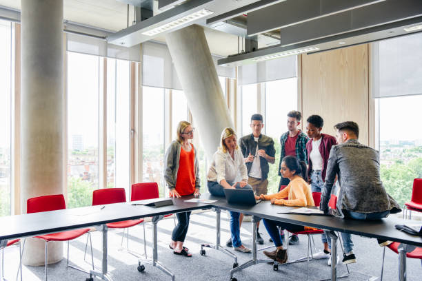 説明する先生のノート パソコンをテーブルの周りの学生のグループ - england talking classroom education ストックフォトと画像