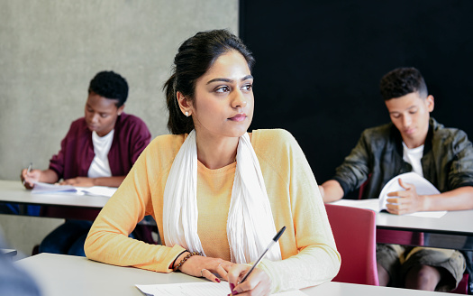 Attractive young Indian woman taking exam in classroom, holding pen and thinking