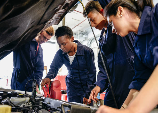 Group of student mechanics working on car engine with hood up Female apprentice attaching electrical jump leads to engine with car bonnet raised, fellow students watching and learning mechanic photos stock pictures, royalty-free photos & images