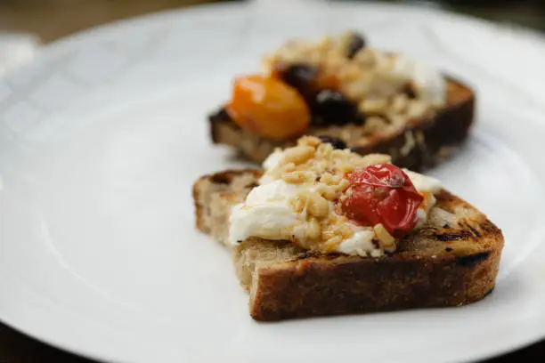 Burrata toasts with cherry tomatoes, olives, ricotta and pinenuts on a white plate