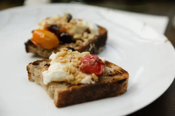 Burrata toasts with cherry tomatoes, olives and pinenuts on a white plate