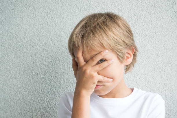 istock Boy covering his face with hand and looking at camera through fingers against grey background 827859348